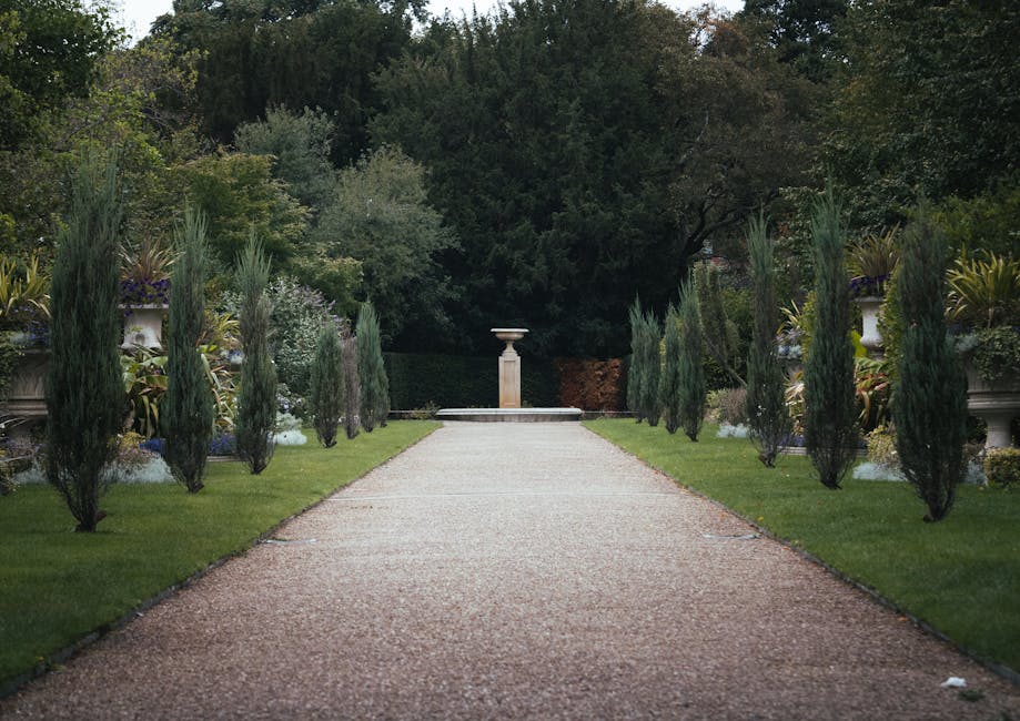 A straight gravel pathway in a landscaped garden, flanked by neatly trimmed grass lawns and a row of evenly spaced tall, slender cypress trees on each side. The pathway leads to a decorative stone ped